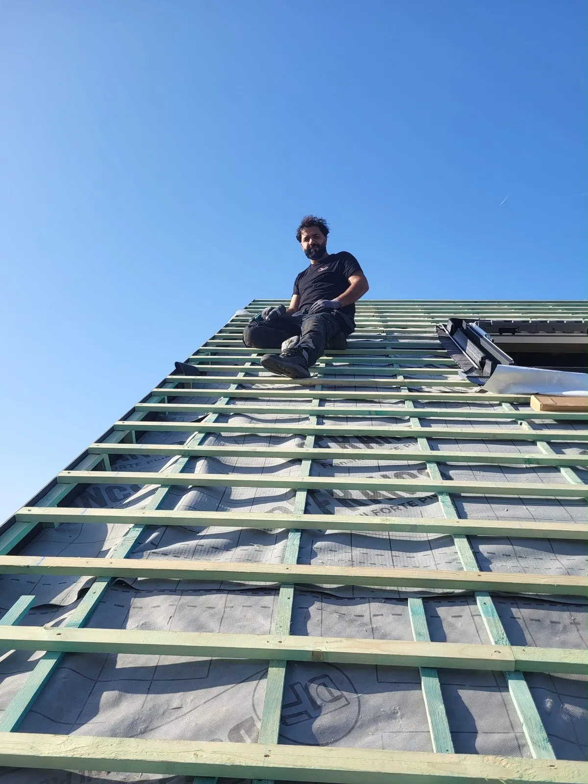 Ciya Ismet op een hellende dakconstructie met onderdak en panlatten, onder een blauwe lucht